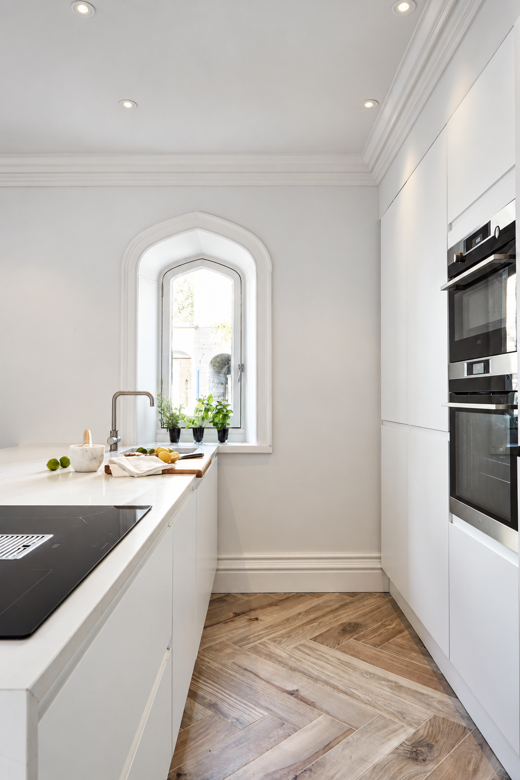White kitchen cabinetry, an island sink and potted herbs beside an arched window at Carter Manor, Wilmslow.