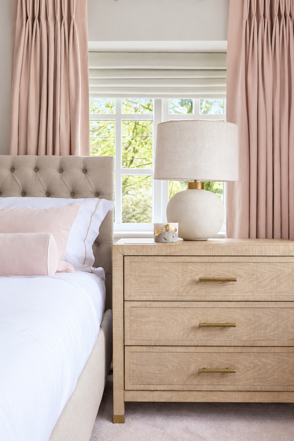 White ceramic bedside lamp and pale drawers beside a buttoned headboard and dusty pink curtains.