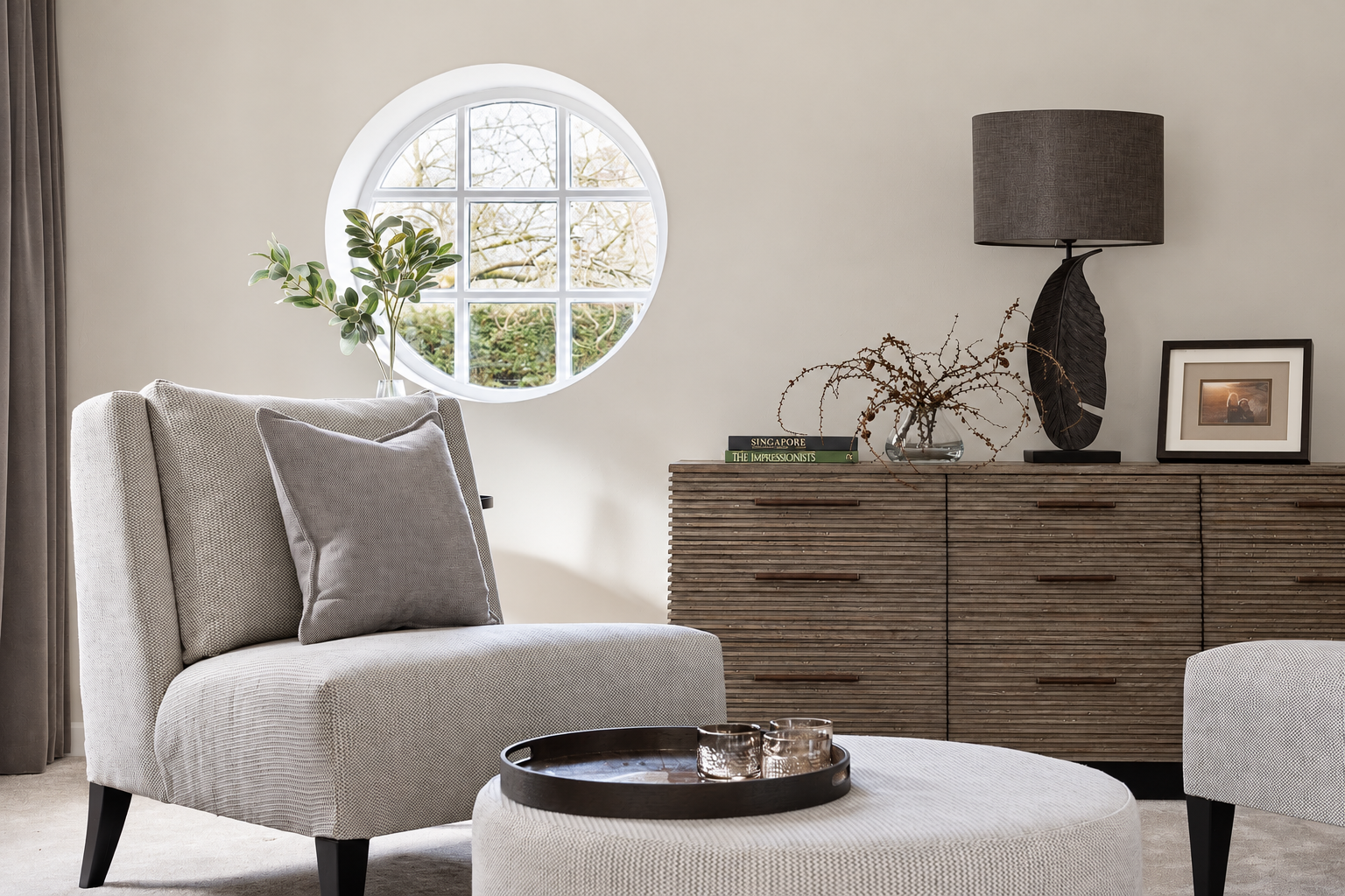 Rounded grey chair beside a circular window, dark drawers and a sculptural table lamp in the living room. At Ivy Cottage, Macclesfield.