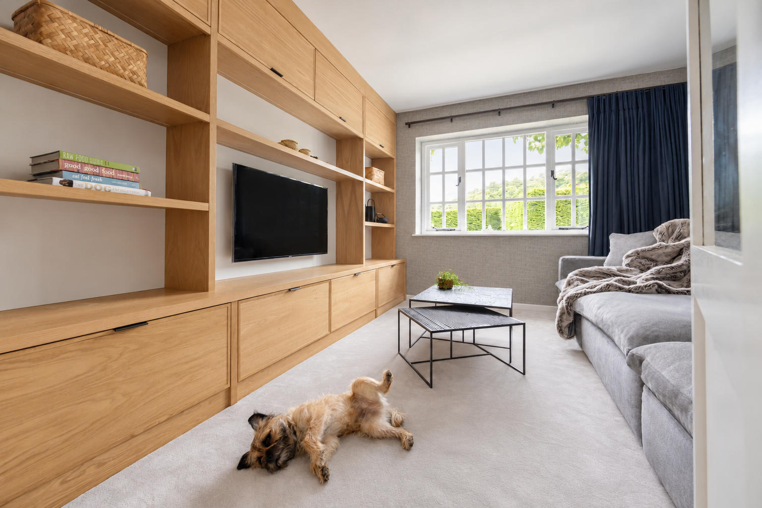 Light oak media cabinetry facing a grey sofa and stone-top nesting tables, with a dog resting on the carpet. At Ivy Cottage, Macclesfield.