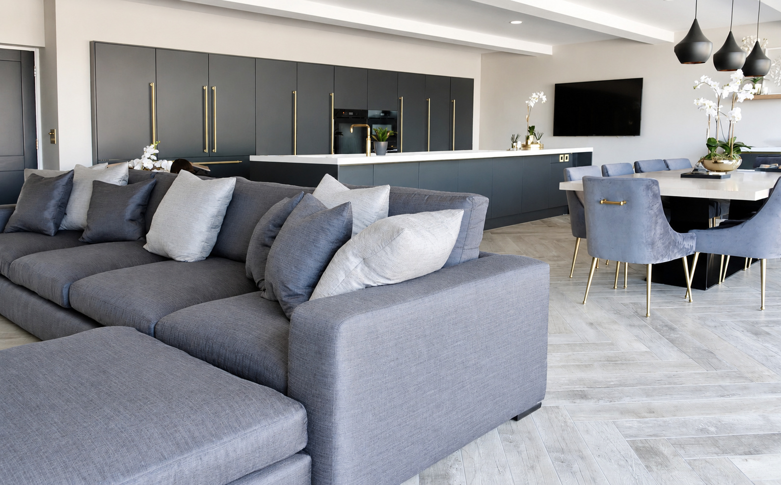 Open-plan kitchen and living room with a blue-grey sofa, dark cabinetry and brass accents at The Upton, Prestbury.