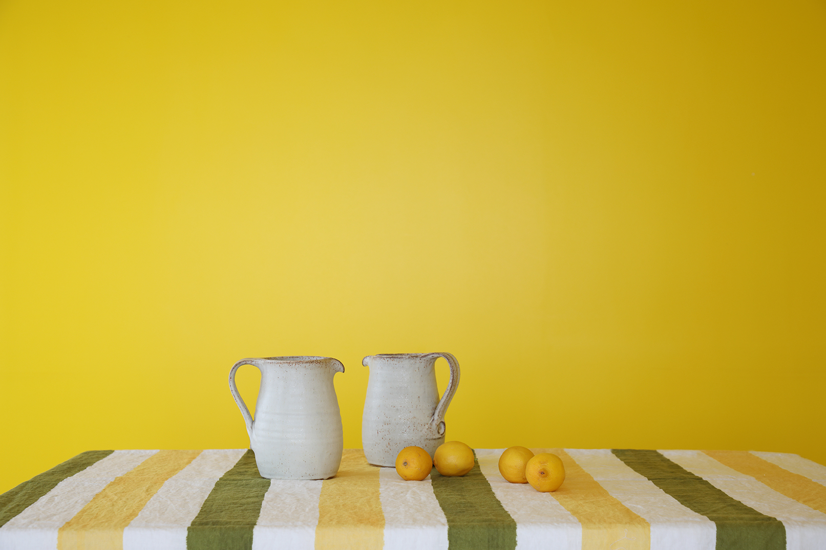 Two cream earthenware jugs and some lemons on a green and yellow striped tablecloth against a bright yellow backdrop