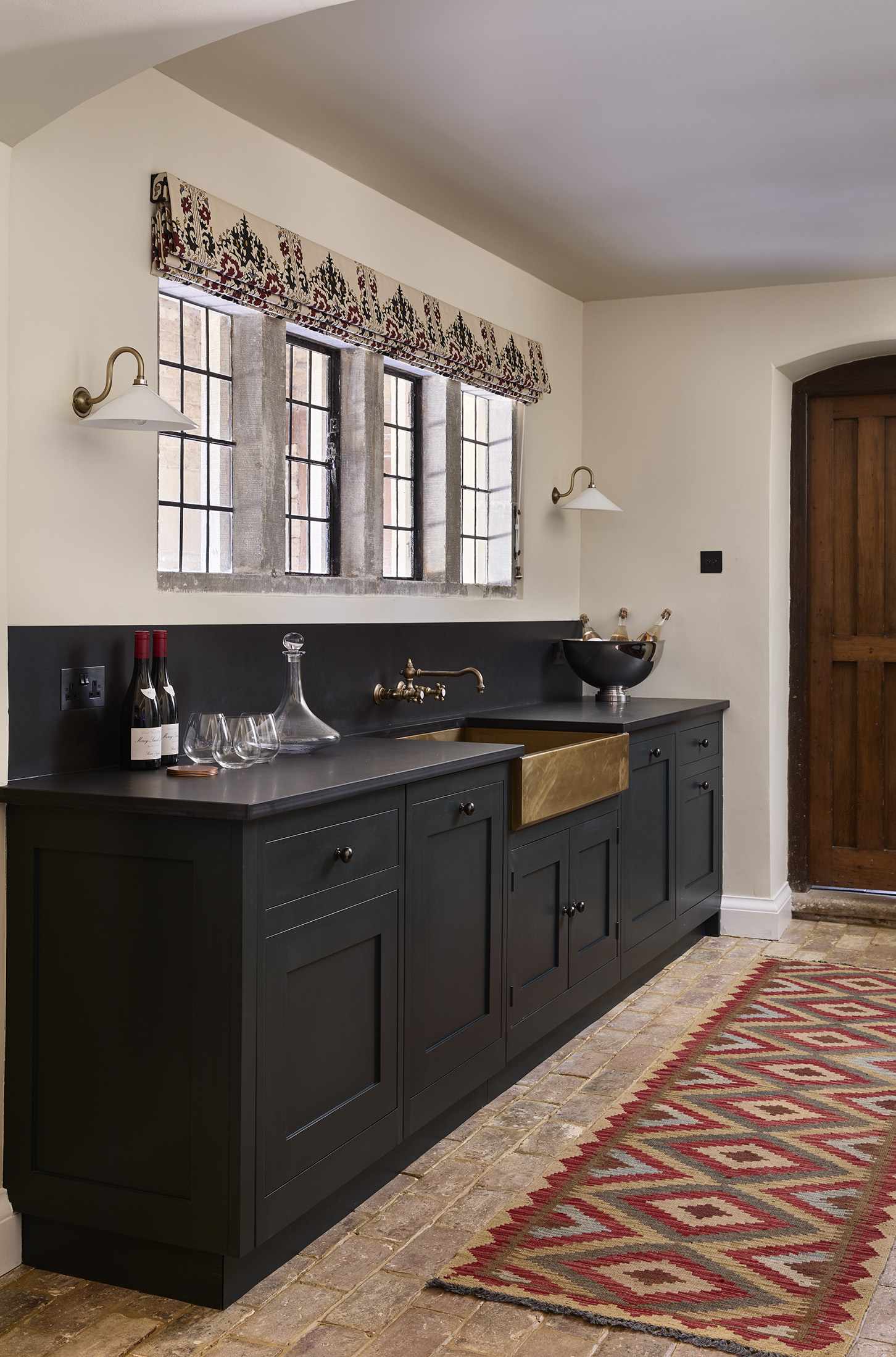 A run of dark grey traditional base cabinets with a large copper rectangular sink, decorative wall lights, & traditional red runner