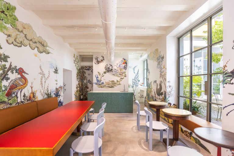 A room with bright bird wallpaper, large bright windows to the right and a red long table with chairs to the left.