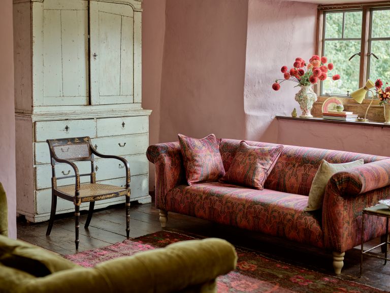 Pink sofa underneath a window in a room with pink walls, wooden floors and rustic chair and cabinet.