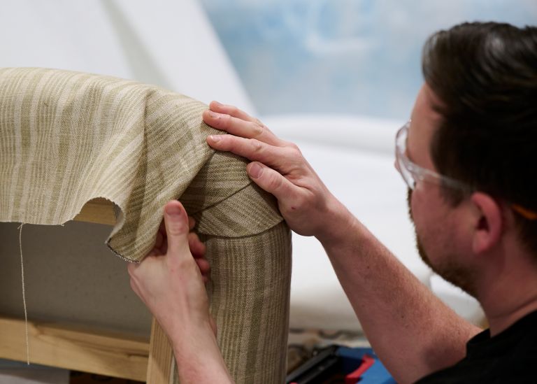 A man wth goggles fitting cream stripy fabric around the corner of a sofa.