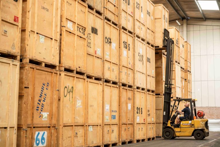 Large storage crates piled high in a warehouse with a cherry picker reaching up to a top one.
