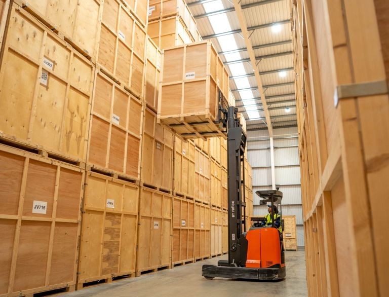 Forklift lifting a large wooden crate onto a stack of large wooden crates in a warehouse.