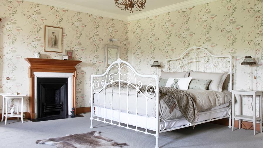 A vintage style cream ironwork bedstead with gentle swirls of passion flowers and buds decorating the wallpaper.