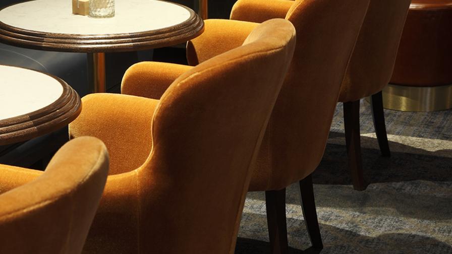 A row of orange velvet armchairs and circular white topped tables with menus on in a bar with moody lighting