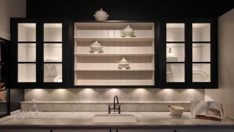 Show kitchen with open shelves and glazed cabinetry above a marble worktop and  basin, with dramatic lighting