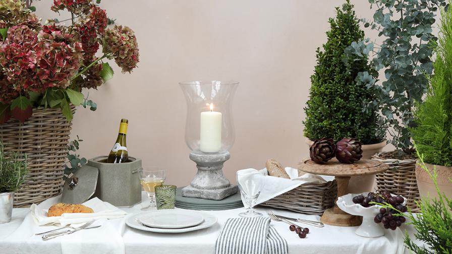 Still life table setting on a white linen tablecloth with a bench in front  surrounded by plants and flowers.