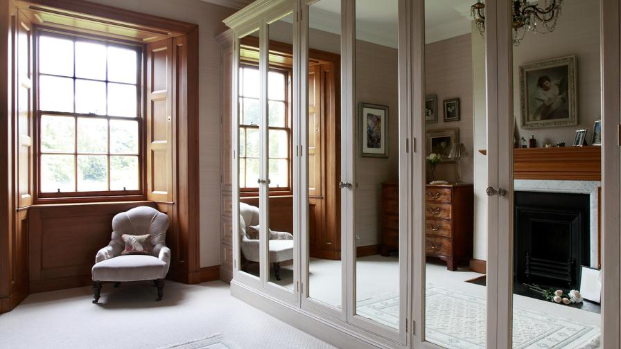 Tall mirror glass door fronts to the bespoke wardrobes have a gilt leaf inset detail and distressed brass rose handles.