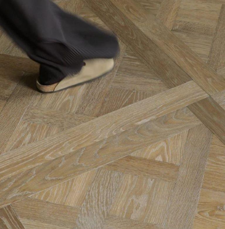 Top down image of person with brown trousers walking on grey-brown Versailles panel parquet flooring.