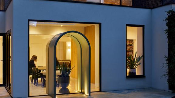 A modern interior space featuring large glass windows and an illuminated arched entryway, creating a warm and inviting atmosphere with visible plants and people inside.