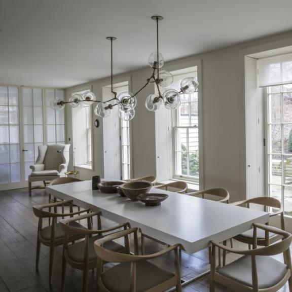 a modern, minimalist dining room with a long white table, wooden chairs, large windows, and stylish pendant lights.